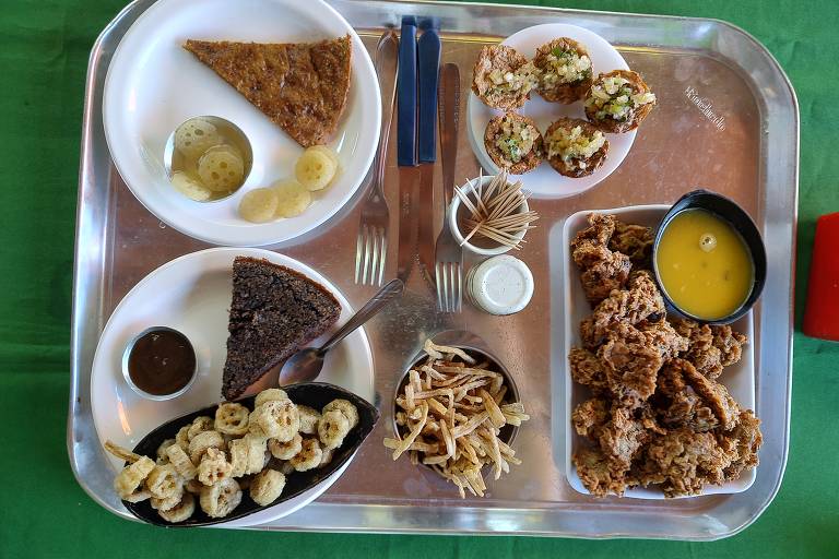 Uma bandeja prateada com diversos pratos de comida. No canto superior esquerdo, há um pedaço de torta e fatias de banana. No canto inferior esquerdo, um prato com bolinhos de banana e um molho escuro. No centro, um prato com um bolo escuro e um pequeno recipiente com molho branco. À direita, há uma porção de frango frito e um recipiente com um líquido amarelo. A bandeja está sobre uma toalha verde.