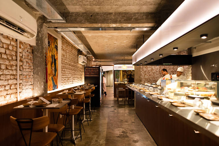 A imagem mostra o interior de um restaurante moderno com paredes de tijolos expostos e um teto de concreto. À esquerda, há mesas e cadeiras de madeira, enquanto à direita, uma bancada exibe pratos de comida. Um chef está visível na área de preparo, e há uma obra de arte colorida na parede. A iluminação é suave e moderna, criando uma atmosfera acolhedora.