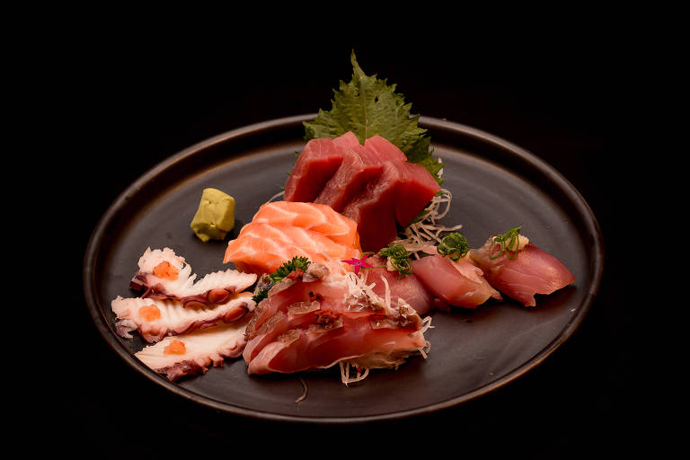 Um prato redondo e escuro contendo uma variedade de sushi. Há fatias de salmão, atum e polvo, decoradas com ervas e wasabi. O fundo é preto, destacando os alimentos.