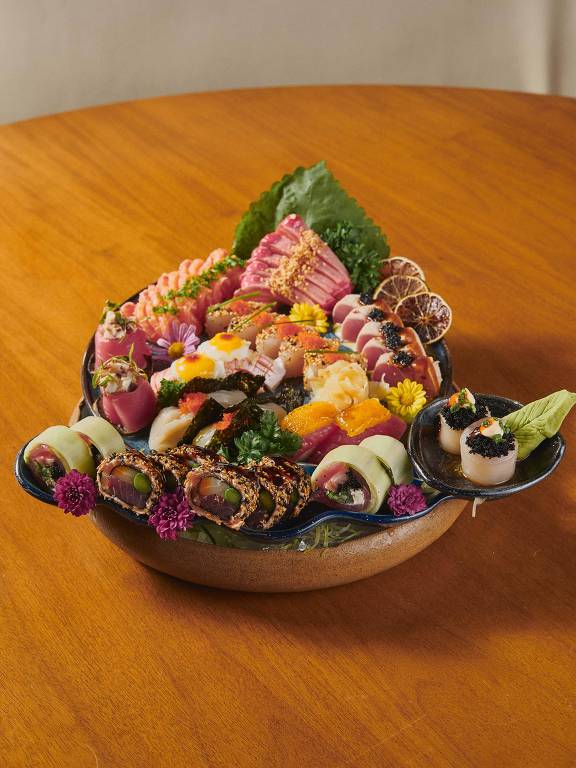 Uma grande travessa de sushi e sashimi disposta artisticamente. A travessa contém uma variedade de peixes, camarões, e outros frutos do mar, decorados com folhas verdes e flores comestíveis. O fundo é uma mesa de madeira clara.