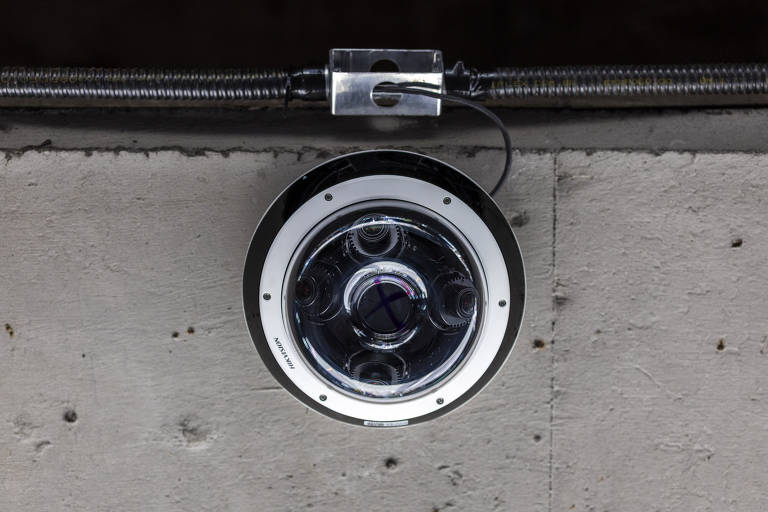 A imagem mostra uma câmera de segurança circular instalada em um teto ou parede. A câmera possui um design moderno, com uma lente central e várias lentes menores ao redor. O fundo é uma superfície de concreto, e há um cabo conectado à câmera, que passa por um suporte metálico