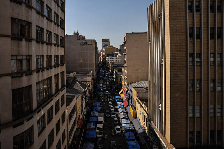 A imagem mostra uma rua movimentada em uma cidade, com prédios altos de ambos os lados. Há uma fila de carros estacionados e barracas de mercado ao longo da rua. O céu está claro e ensolarado, e várias bandeiras estão visíveis nas barracas. A perspectiva é de um ponto elevado, proporcionando uma visão ampla da rua.