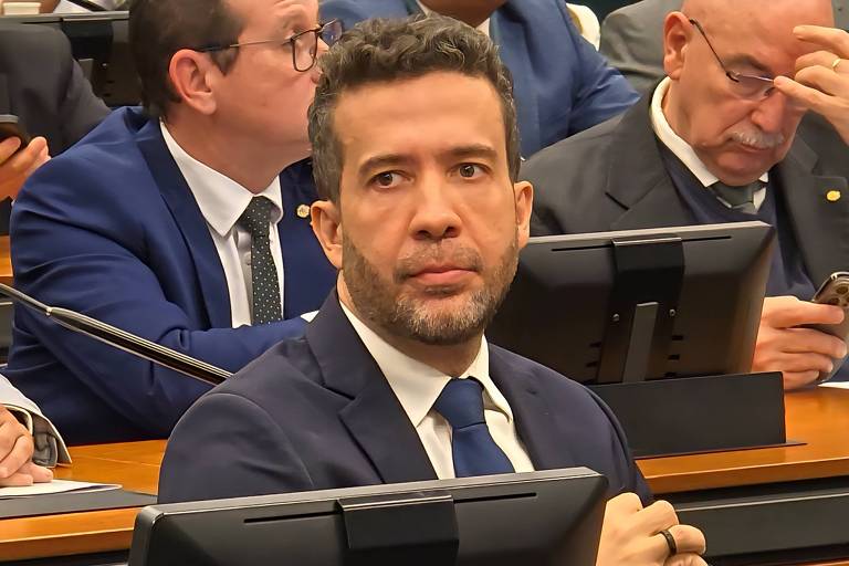 L'image montre un homme aux cheveux courts et à la barbe, vêtu d'un costume sombre et d'une cravate. Il est assis à une table lors d'une séance législative, l'air sérieux. À l'arrière-plan, d'autres personnes sont présentes, certaines regardant leur téléphone, d'autres attentifs. Le décor semble être une salle de réunion ou une séance plénière.