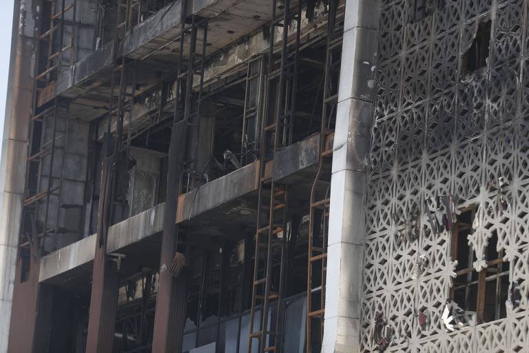 A imagem mostra a fachada de um edifício danificado, com várias janelas quebradas e estruturas metálicas expostas. A parede do edifício apresenta um padrão de revestimento, e há sinais de incêndio, como áreas carbonizadas e desmoronadas. A cena transmite uma sensação de abandono e destruição.