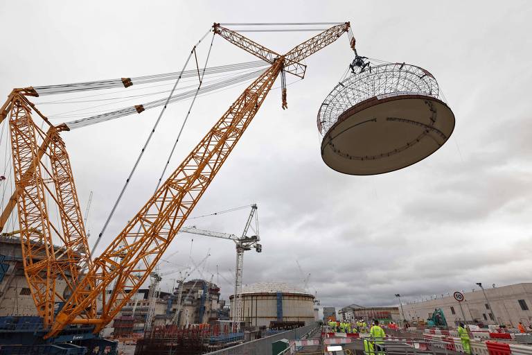 A imagem mostra um grande guindaste amarelo levantando uma estrutura circular em um canteiro de obras. Ao fundo, há outros guindastes e partes de uma construção em andamento. O céu está nublado, e várias pessoas vestindo coletes de segurança estão visíveis na área de trabalho.