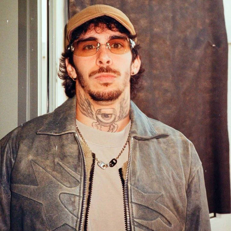 Um homem está posando para a foto, usando uma jaqueta cinza com detalhes escuros e uma camiseta clara. Ele tem cabelo escuro e encaracolado, e usa óculos de sol e um boné. O homem tem várias tatuagens visíveis no pescoço e no rosto. Ao fundo, há uma cortina escura que parece ser de tecido. A iluminação é suave, destacando o rosto do homem.