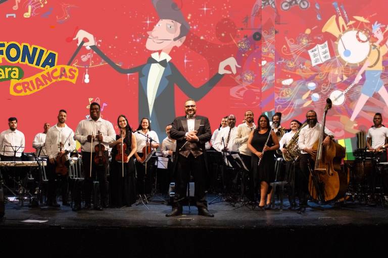 A imagem mostra uma orquestra em um palco, com músicos de pé e sentados, todos vestidos de forma formal, em sua maioria com camisas brancas. No centro, um maestro está de pé, usando um terno escuro e segurando uma batuta. Ao fundo, há um grande painel colorido com a inscrição 'SINFONIA para CRIANÇAS' e ilustrações animadas. A cena transmite um ambiente festivo e musical.