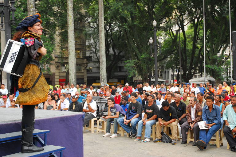 A imagem mostra uma apresentação de rua em uma praça, onde um artista vestido de forma colorida e com um acordeão está se apresentando em um palco. O artista está de perfil, com uma expressão animada, enquanto uma grande plateia observa atentamente. A audiência é composta principalmente por homens e mulheres de diversas idades, sentados em cadeiras plásticas. Ao fundo, há árvores e prédios, indicando um ambiente urbano.