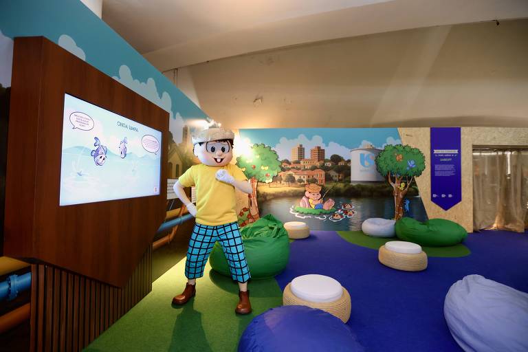 A imagem mostra um espaço infantil decorado com um personagem de desenho animado, que está vestido com uma camiseta amarela e calças xadrez azuis. O personagem está posando em frente a uma tela que exibe uma animação. Ao fundo, há uma parede decorada com ilustrações de uma cidade e árvores, além de almofadas coloridas dispostas pelo chão.