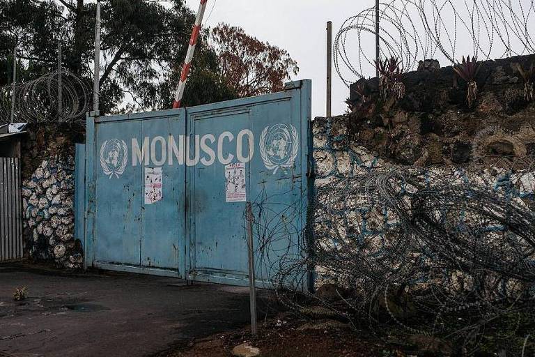 L'immagine mostra un cancello blu con il logo della MONUSCO (Missione delle Nazioni Unite nella Repubblica Democratica del Congo). Il cancello è circondato da un muro di pietra e filo spinato. Intorno c'è vegetazione e il cielo appare nuvoloso.