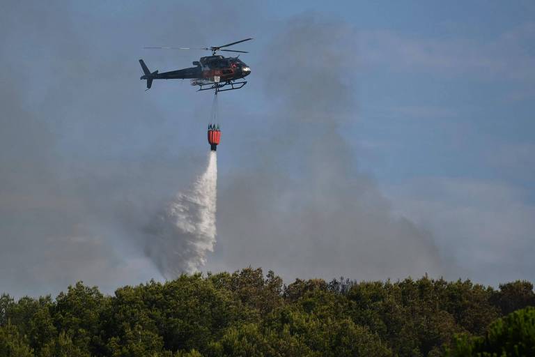 Um helicóptero está sobrevoando uma área com fumaça, despejando água ou um agente extintor de um balde suspenso. A vegetação verde está visível abaixo, contrastando com a fumaça que se eleva no ar.