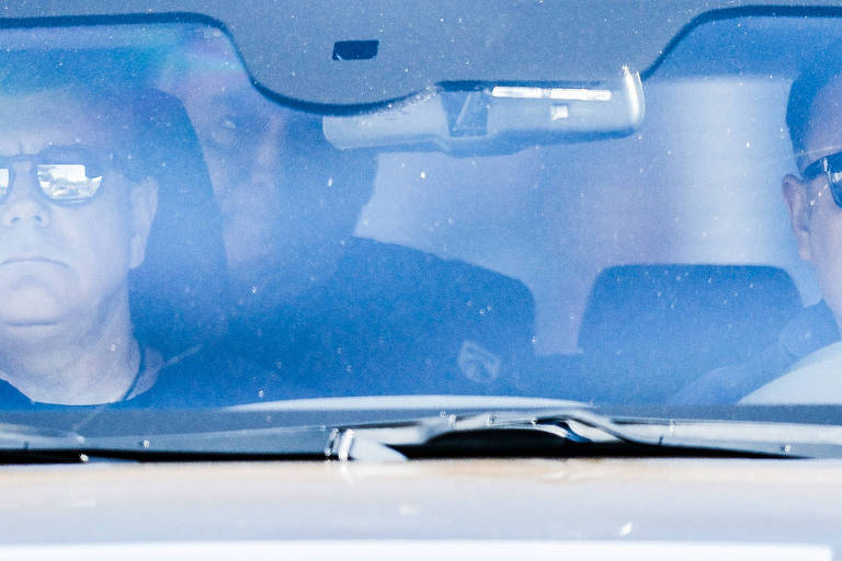 A imagem mostra três homens dentro de um carro, visíveis através do para-brisa. Dois deles estão na frente, usando óculos escuros, enquanto um terceiro homem está parcialmente visível atrás deles. O interior do carro é visível, mas a imagem tem um leve reflexo e sujeira no vidro, dificultando a visualização clara dos rostos.