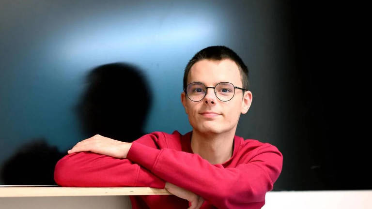 Um jovem está posando para a foto, com os braços apoiados em uma superfície. Ele usa um suéter vermelho e óculos. O fundo é escuro, com uma leve iluminação que destaca seu rosto e cria uma sombra ao lado.