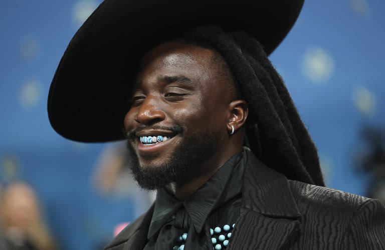Um homem sorridente usa um grande chapéu preto e uma roupa escura. Ele tem dreadlocks e dentes com aparelho dental azul. O fundo é desfocado, com algumas luzes e pessoas ao longe.