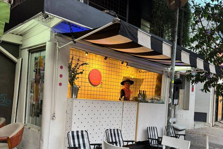 A imagem mostra uma pequena cafeteria localizada na calçada. A estrutura é branca com uma fachada que possui uma grade de azulejos e uma grande janela. Um funcionário está atrás do balcão, e há mesas e cadeiras dispostas do lado de fora, algumas com listras pretas e brancas. O ambiente é iluminado e há plantas decorativas ao redor.