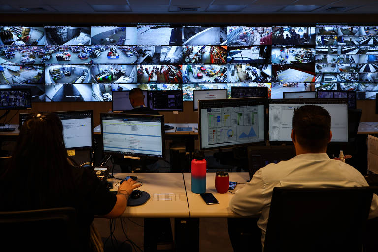 A imagem mostra um centro de monitoramento com várias telas exibindo imagens de câmeras de segurança. Duas pessoas estão sentadas em frente a computadores, uma delas é uma mulher com cabelo longo e a outra é um homem com cabelo curto. As telas na parede mostram diferentes ângulos de câmeras, algumas com imagens de pessoas e veículos. O ambiente é bem iluminado e organizado.