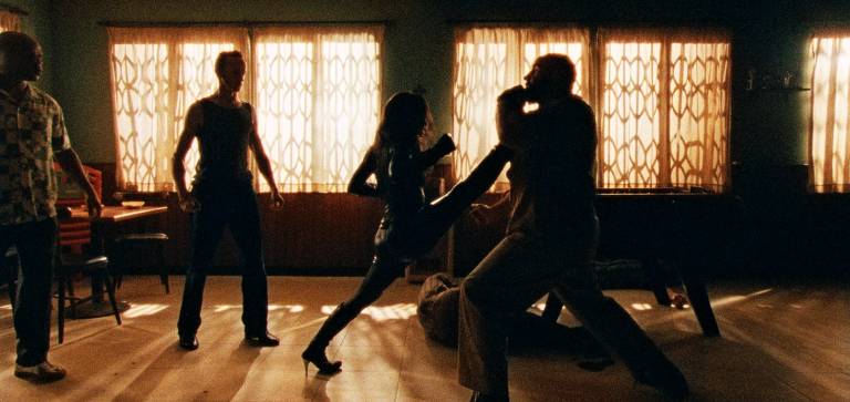 A imagem mostra uma cena de luta em um ambiente interno com luz suave. Quatro pessoas estão presentes: duas estão em uma posição de combate, uma mulher está chutando um homem, enquanto os outros dois observam. As janelas ao fundo têm cortinas que filtram a luz, criando um efeito dramático na cena.