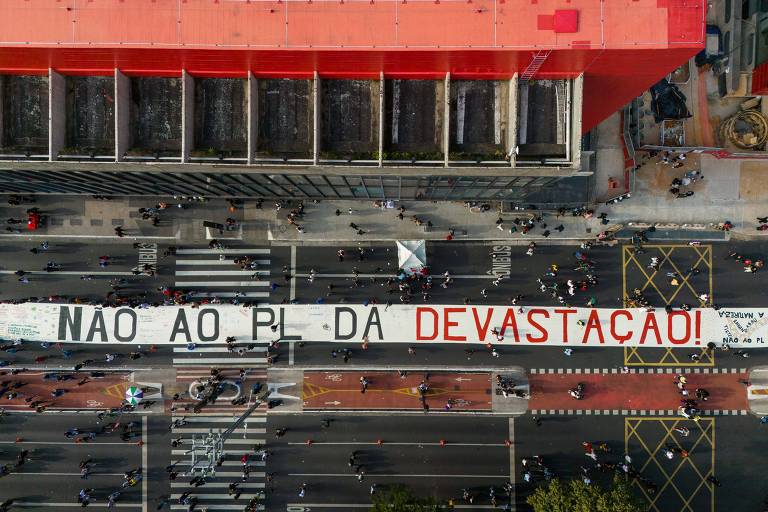 A imagem mostra uma grande faixa estendida sobre uma rua, com a inscrição em letras grandes: 'NÃO AO PL DA DEVASTAÇÃO'. A faixa ocupa a largura da via, e há várias pessoas caminhando ao redor. O cenário é urbano, com edifícios ao fundo e uma área de calçada visível. A faixa é predominantemente branca com letras vermelhas. A imagem é capturada de uma perspectiva aérea, mostrando a movimentação na rua.
