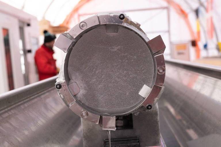 A imagem mostra a parte frontal de um equipamento cilíndrico, possivelmente um tubo ou um cilindro, com uma superfície metálica e detalhes em prata. O fundo é desfocado, mas é possível ver uma estrutura de tenda ou abrigo, com uma pessoa vestindo um casaco vermelho ao fundo. O cilindro parece estar em um ambiente de pesquisa ou laboratório.