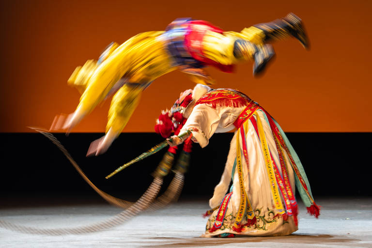 A imagem mostra dois dançarinos em uma apresentação acrobática. Um dançarino, vestido com um traje amarelo e azul, está saltando no ar, enquanto o outro, vestido com um traje tradicional colorido, se inclina para frente, segurando um bastão. O fundo é de uma cor laranja suave, destacando a ação dos dançarinos.
