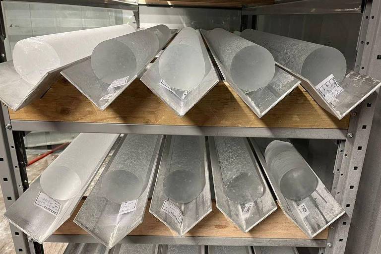 A imagem mostra uma estante metálica com várias peças de metal dispostas em camadas. As peças incluem tubos cilíndricos e perfis em forma de L, todos com uma superfície prateada e etiquetas visíveis. A estante é organizada e as peças estão alinhadas de forma ordenada, com algumas peças mais longas e outras mais curtas.