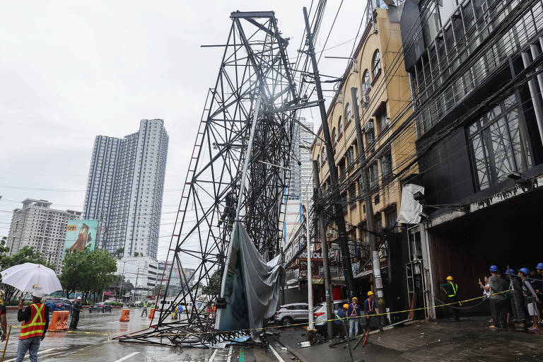 A imagem mostra uma estrutura de metal, possivelmente um outdoor, caída em uma área urbana. A estrutura está inclinada e danificada, com fios elétricos expostos. Ao fundo, há prédios altos e uma rua com veículos. Algumas pessoas estão na cena, algumas usando capacetes e coletes de segurança, enquanto outras seguram um guarda-chuva. O céu está nublado, indicando um clima chuvoso.