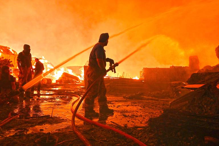 A imagem mostra bombeiros lutando contra um incêndio intenso. Eles estão usando mangueiras para combater as chamas que consomem uma área ao fundo, envolta em fumaça e luz laranja. O solo está molhado, refletindo a luz do fogo e a água das mangueiras. A cena transmite um senso de urgência e perigo.