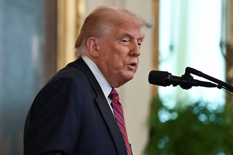 Um homem está falando em um microfone em um ambiente formal. Ele usa um terno escuro e uma gravata vermelha. Ao fundo, há uma decoração que sugere um ambiente de evento, com plantas e uma parede decorada. O homem parece estar em um momento de discurso ou apresentação.
