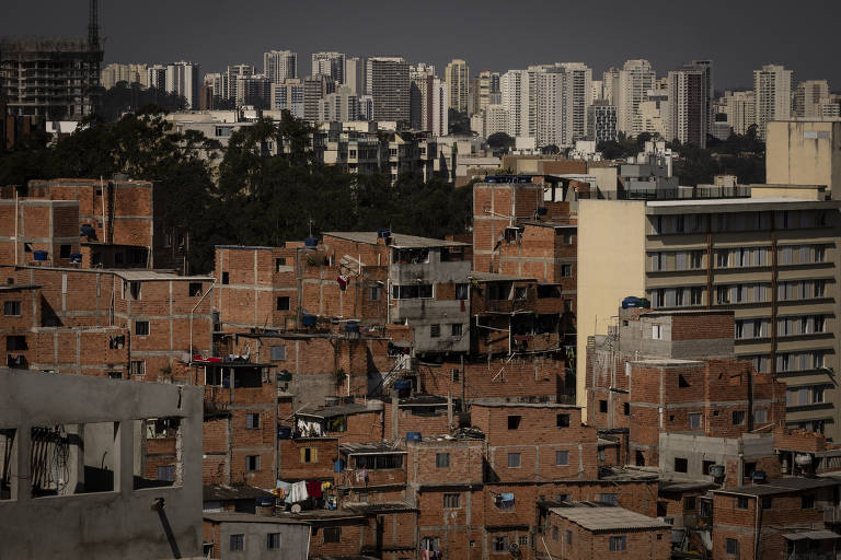 A imagem mostra uma vista de uma área urbana com prédios de apartamentos altos ao fundo, enquanto em primeiro plano há uma série de casas de tijolos, típicas de uma comunidade de baixa renda. As casas são pequenas e estão próximas umas das outras, com algumas varandas e roupas estendidas. O contraste entre as construções simples e os edifícios modernos ao fundo é evidente, refletindo a desigualdade urbana.
