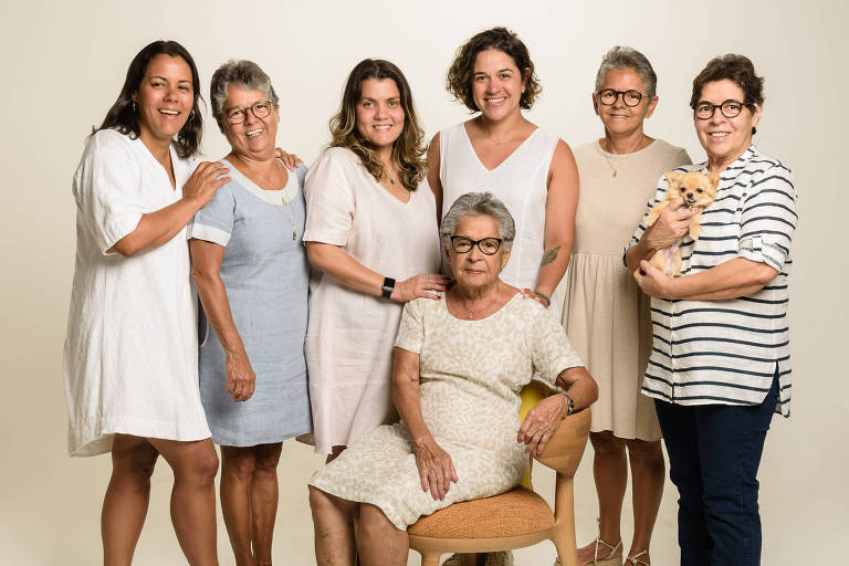 A imagem mostra um grupo de sete mulheres posando juntas. Elas estão em um fundo claro e neutro. A mulher sentada no centro usa um vestido claro e segura a mão em uma posição relaxada. As outras mulheres estão em pé ao redor dela, algumas usando vestidos e outras blusas e calças. Uma das mulheres em pé segura um gato laranja. Todas parecem sorrir e estão em um ambiente descontraído.