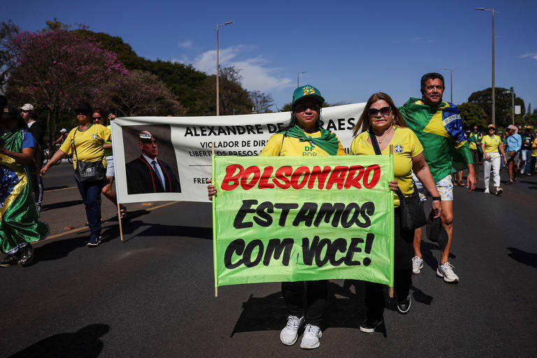 A imagem mostra uma manifestação na rua, onde várias pessoas estão marchando. No primeiro plano, uma mulher segura um cartaz verde com letras vermelhas que diz 'BOLSONARO ESTAMOS COM VOCÊ!'. Ao fundo, há uma faixa com o nome 'ALEXANDRE DE MORAES' e outras palavras relacionadas a direitos. As pessoas estão vestidas com camisetas amarelas e verdes, representando as cores da bandeira do Brasil, e algumas estão segurando bandeiras brasileiras. O ambiente é urbano, com árvores e postes ao fundo.