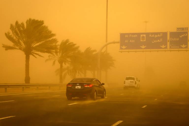 A imagem mostra uma estrada em meio a uma tempestade de areia, com visibilidade reduzida. À esquerda, há palmeiras balançando ao vento, e à direita, um sinal de trânsito com indicações em árabe e inglês. Dois carros estão visíveis na estrada, um preto e um branco, ambos se movendo em direção à câmera.