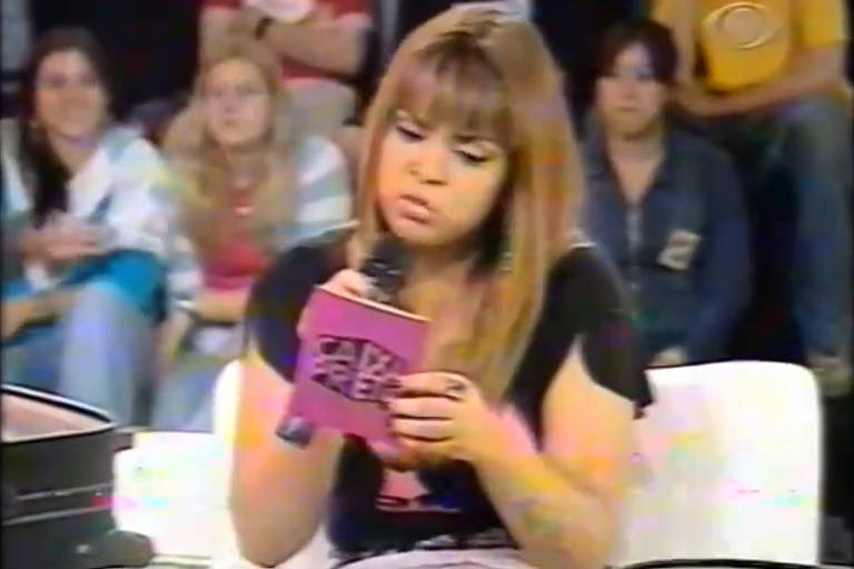 Uma jovem mulher com cabelo longo e liso, usando uma camiseta preta, está sentada em um sofá segurando um microfone e um caderno rosa. Ela parece concentrada enquanto lê algo. Ao fundo, há um público, com várias pessoas assistindo ao programa, algumas sorrindo e outras atentas.