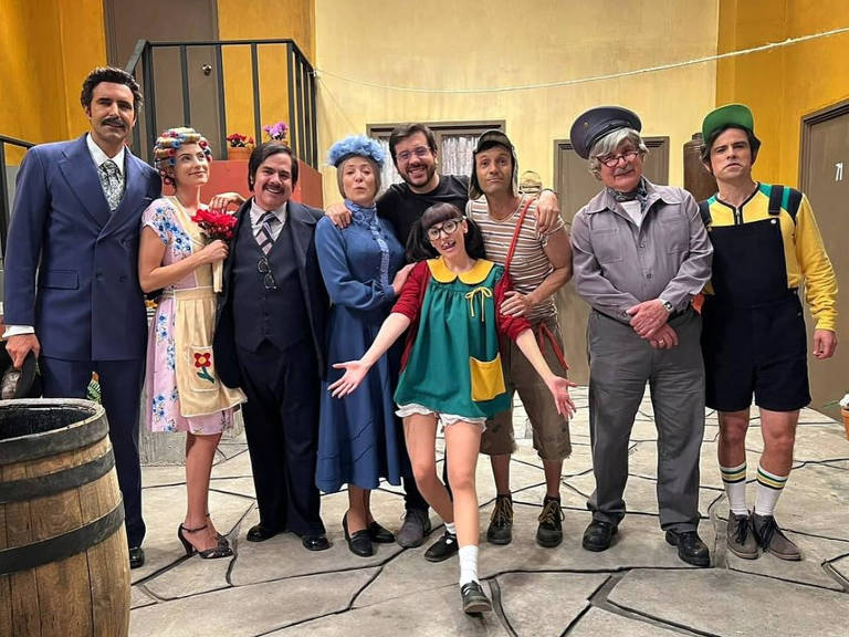 Um grupo de nove pessoas posando para a foto em um cenário que parece ser de um estúdio de televisão. As pessoas estão vestidas com roupas coloridas e características de personagens de comédia. Há um barril de madeira à esquerda e as paredes são de um tom amarelo. O grupo inclui homens e mulheres, com expressões alegres e descontraídas, e uma das mulheres está em destaque na frente, fazendo uma pose divertida.