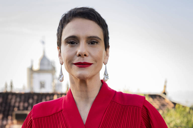 Uma mulher está posando em um ambiente externo, vestindo um vestido vermelho com detalhes plissados e mangas largas. Ela tem cabelo curto e usa brincos grandes. Ao fundo, há uma construção com uma cúpula visível e um céu claro.