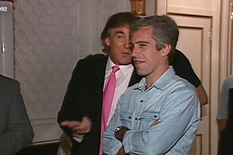 A imagem mostra um grupo de homens em um ambiente interno, possivelmente em um evento social. Um homem à esquerda, com cabelo laranja e terno escuro, está falando e gesticulando. Ao seu lado, um homem com cabelo grisalho e camisa jeans está sorrindo e com os braços cruzados. Outros homens estão visíveis ao fundo, mas não estão em foco. O ambiente tem paredes decoradas e uma iluminação suave.