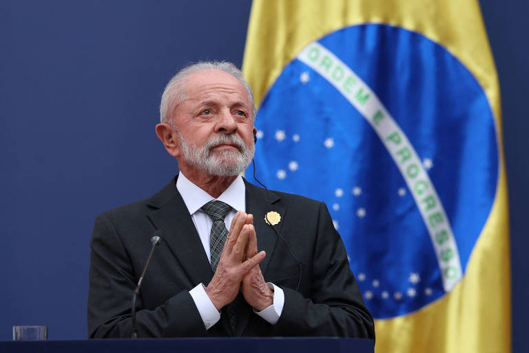 Um homem de cabelos grisalhos e barba, vestido com um terno escuro e gravata, está em pé com as mãos unidas em um gesto de agradecimento ou oração. Ao fundo, há uma bandeira do Brasil, com as cores verde, amarelo e azul, visível em destaque. O homem parece estar em um evento formal, possivelmente fazendo um discurso ou declaração.