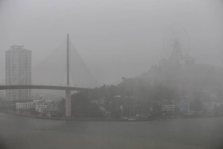 A imagem mostra uma vista de uma cidade em um dia nublado e chuvoso. Há uma ponte com um design moderno que se estende sobre um rio. Ao fundo, é possível ver um edifício alto e uma roda-gigante, ambos parcialmente ocultos pela névoa. A visibilidade é baixa devido à neblina, e a cena transmite uma atmosfera sombria e melancólica.