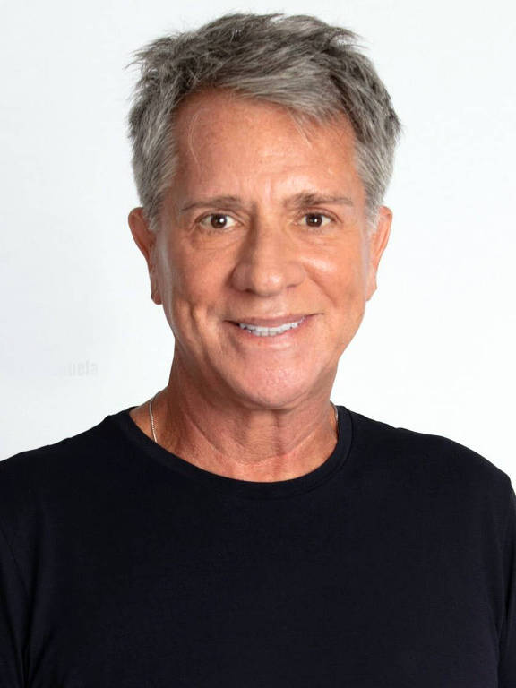 Um homem sorridente está posando para a foto. Ele tem cabelo grisalho e usa uma camiseta preta. O fundo é claro e neutro, destacando a figura do homem.