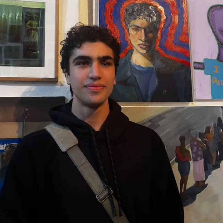 Um jovem com cabelo cacheado e escuro está posando em frente a uma parede de galeria de arte. Ele usa um moletom preto e uma bolsa cruzada. Ao fundo, há várias obras de arte, incluindo um retrato de um homem com cabelo cacheado e uma camisa azul, cercado por um fundo vermelho. Outras pinturas e fotografias estão visíveis na parede.