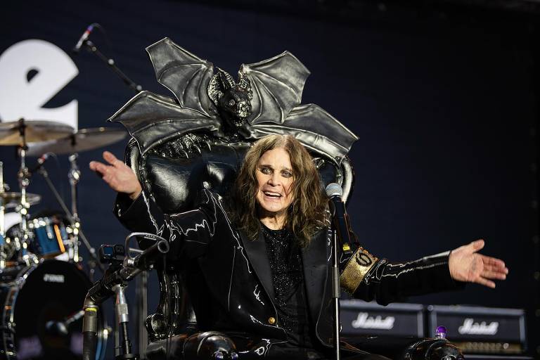 Um artista está sentado em uma cadeira grande e ornamentada, com um design que inclui um morcego em cima. Ele está usando um traje preto e tem cabelo longo e solto. O artista está sorrindo e gesticulando com as mãos, enquanto um microfone está posicionado à sua frente. Ao fundo, há instrumentos musicais e equipamentos de som, sugerindo que ele está em um palco durante uma apresentação.