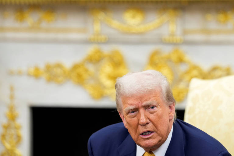 The image shows a man with blond hair and fair skin, sitting in a formal setting. He's wearing a dark suit and a white shirt. In the background, there's a wall decorated with gold accents and a light-colored table or sofa. The man appears to be in conversation or paying attention.