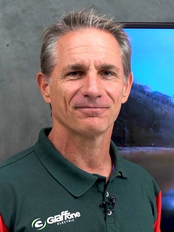 Um homem está posando para a foto, usando uma camisa polo verde com detalhes em vermelho. Ele tem cabelo grisalho e está em um ambiente interno, com uma parede de tijolos ao fundo e uma tela ao lado. O homem parece estar sorrindo levemente e está de pé, com os braços ao lado do corpo.