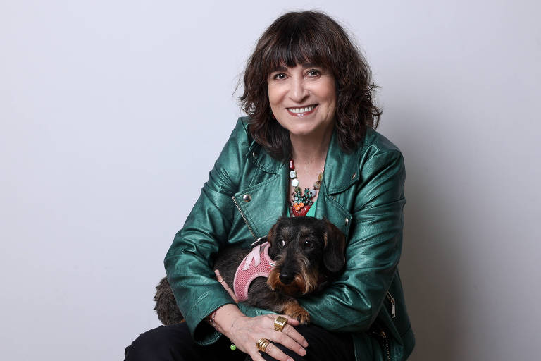 Uma mulher está sentada, sorrindo, com um cachorro no colo. Ela usa uma jaqueta de couro verde e tem cabelo castanho escuro e ondulado. O cachorro é pequeno, com pelagem escura e usa um colete rosa. O fundo da imagem é claro e neutro, destacando a mulher e o cachorro.