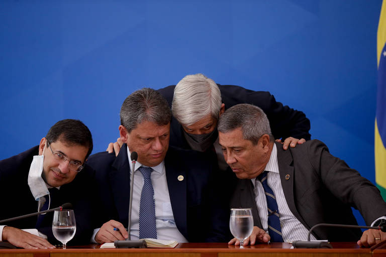 Four men are gathered at a table, leaning over to discuss something. They are dressed formally, with ties and shirts. One of the men wears a face mask. In the background is a Brazilian flag. The table contains glasses of water and a notebook.