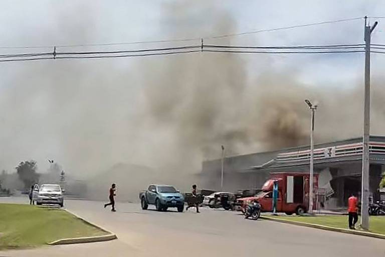 A imagem mostra uma cena de um incêndio em uma área urbana. Há uma grande quantidade de fumaça escura subindo no céu, proveniente de um edifício à direita. Algumas pessoas estão visíveis na rua, com um homem atravessando a via. Vários veículos estão estacionados e em movimento, incluindo um carro azul e um caminhão vermelho. O céu está parcialmente nublado e a vegetação é visível ao fundo.