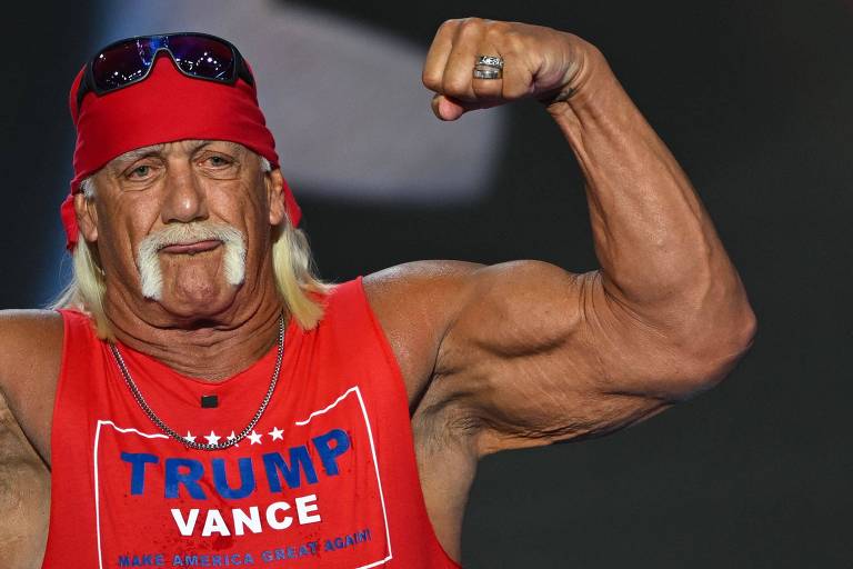 Um homem musculoso está posando com um braço flexionado, mostrando seus músculos. Ele usa uma bandana vermelha e óculos escuros. Sua camiseta é vermelha com a palavra 'TRUMP' e 'VANCE' estampadas. O fundo é desfocado, sugerindo um evento ou apresentação.