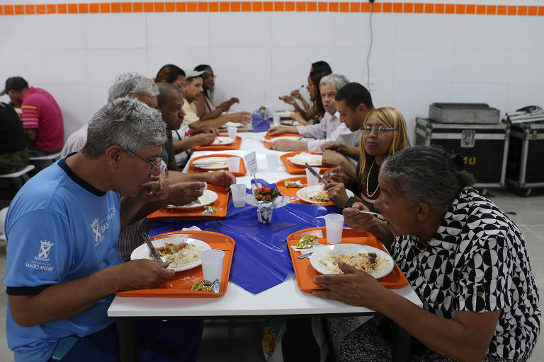 Um grupo de pessoas está sentado em uma mesa longa, compartilhando uma refeição. Cada pessoa tem um prato de comida e um copo. A mesa está coberta com uma toalha azul e há bandejas laranjas com comida. As pessoas parecem estar conversando e se servindo. O ambiente é simples, com paredes brancas e detalhes laranja.