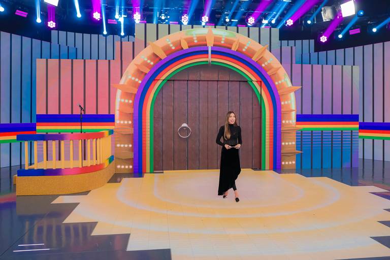 A imagem mostra um cenário de programa de televisão com um arco colorido em tons de rosa, azul, verde e amarelo. No centro, há uma porta marrom com um anel. À esquerda, há uma estrutura em forma de palco com degraus e uma barra colorida. Uma apresentadora está em pé no centro do palco, vestindo uma roupa preta. O fundo é iluminado com luzes coloridas.