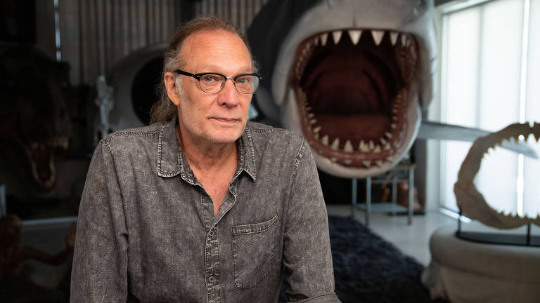 Um homem de cabelos longos e óculos está sentado em primeiro plano, vestindo uma camisa de jeans. Ao fundo, há uma grande escultura de um tubarão com a boca aberta, mostrando dentes afiados. O ambiente parece ser um estúdio ou uma sala de exposições, com luz natural entrando pelas janelas.